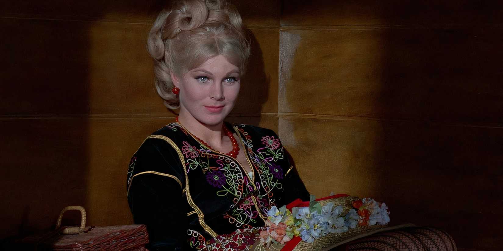 a blonde woman sits smiling enigmatically while wearing a dark floral top