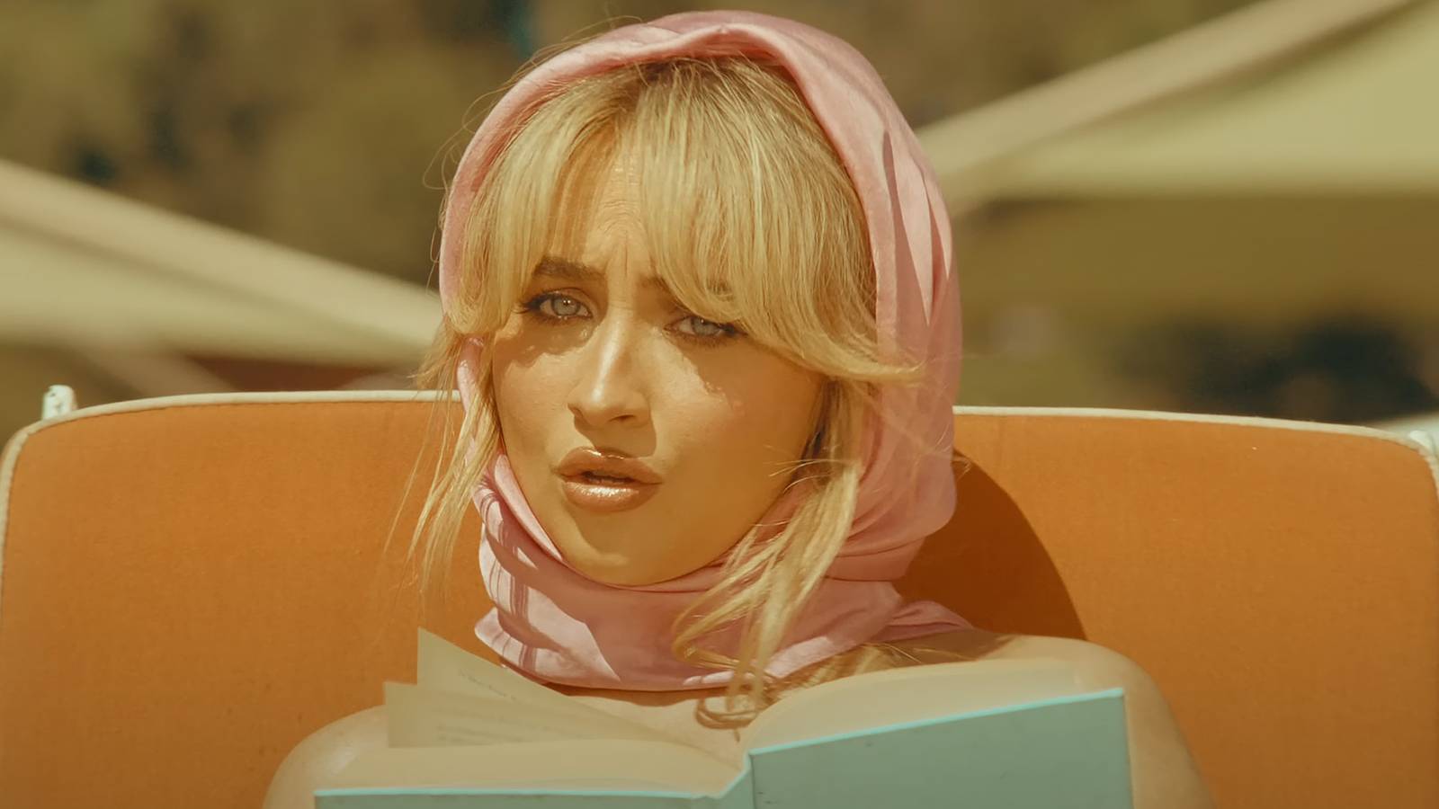 Sabrina Carpenter wearing head scarf reading book on beach lounger in Espresso music video
