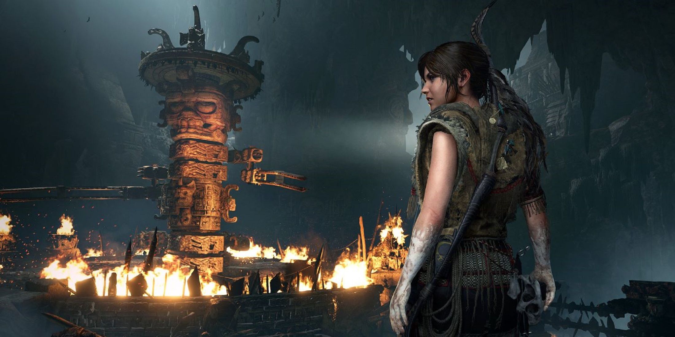 Lara Croft stands in a dark cave, observing a large, burning ancient mechanism in Shadow of the Tomb Raider