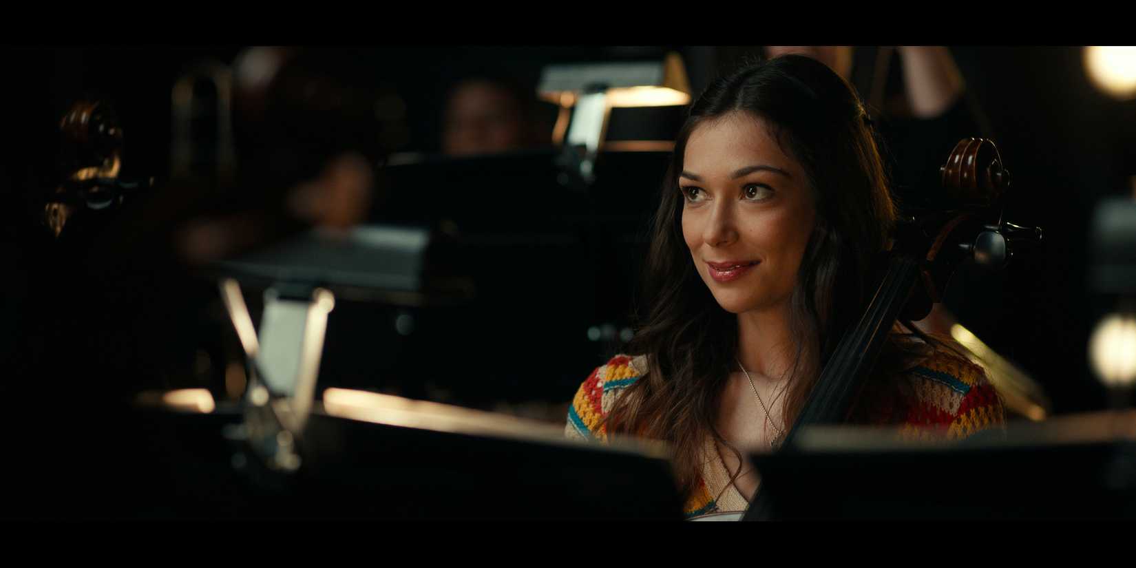 A still from Side Quest episode "Fugue" with a woman sitting in an orchestra pit holding a cello and smiling