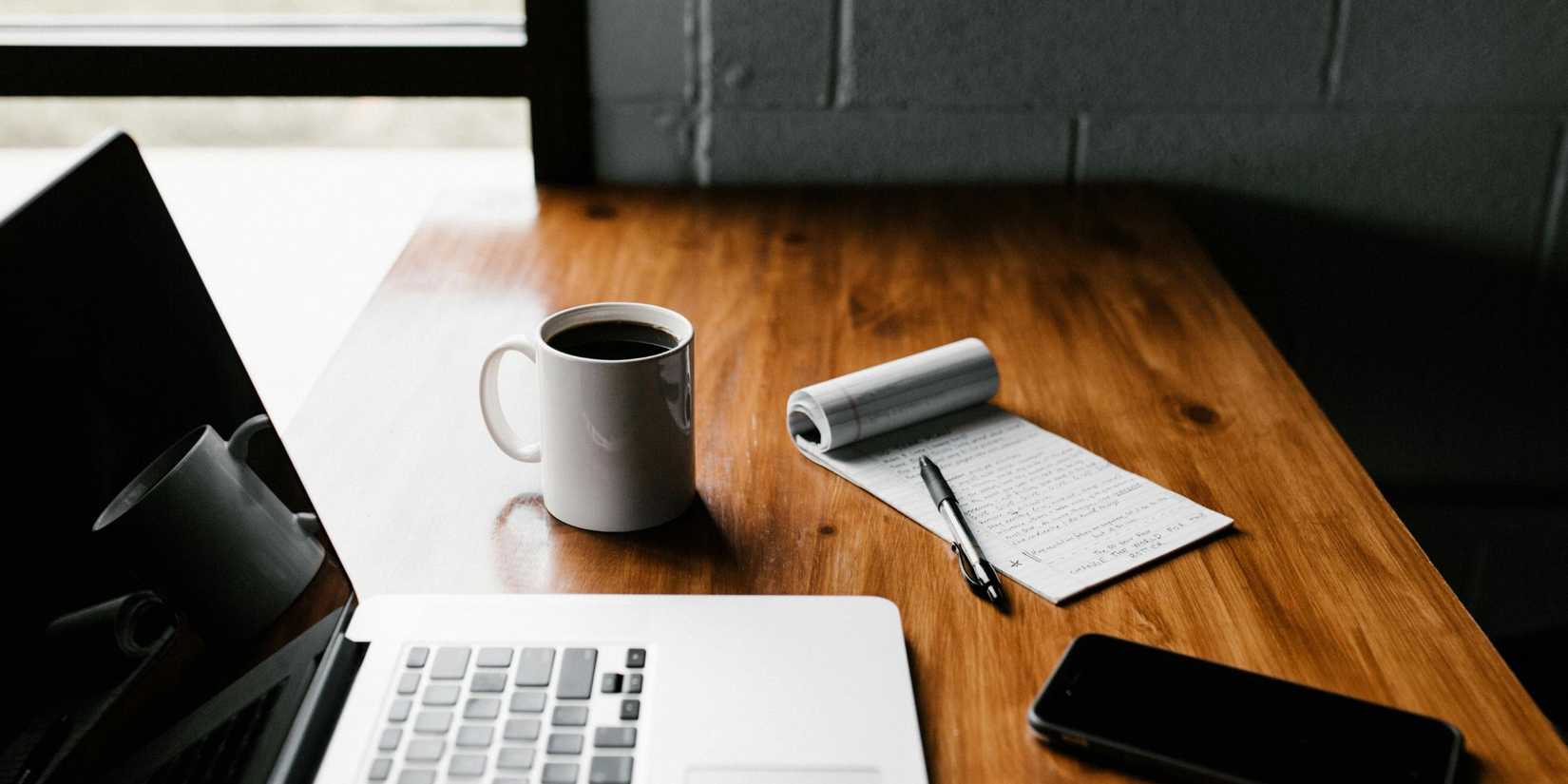 Image of a laptop on a desk with some coffee and a notepad and a mobile.