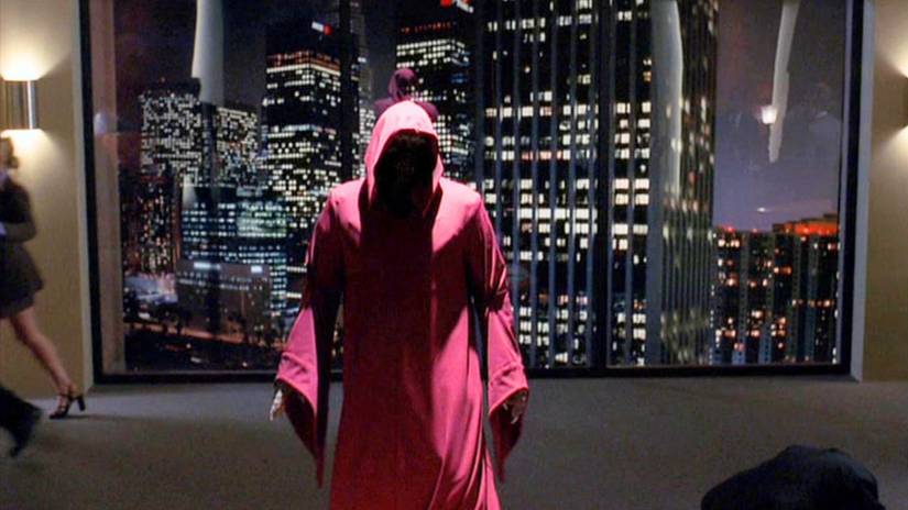 A shadowy figured in a red hooded robe stands in front of a window showing the Los Angeles skyline in Angel
