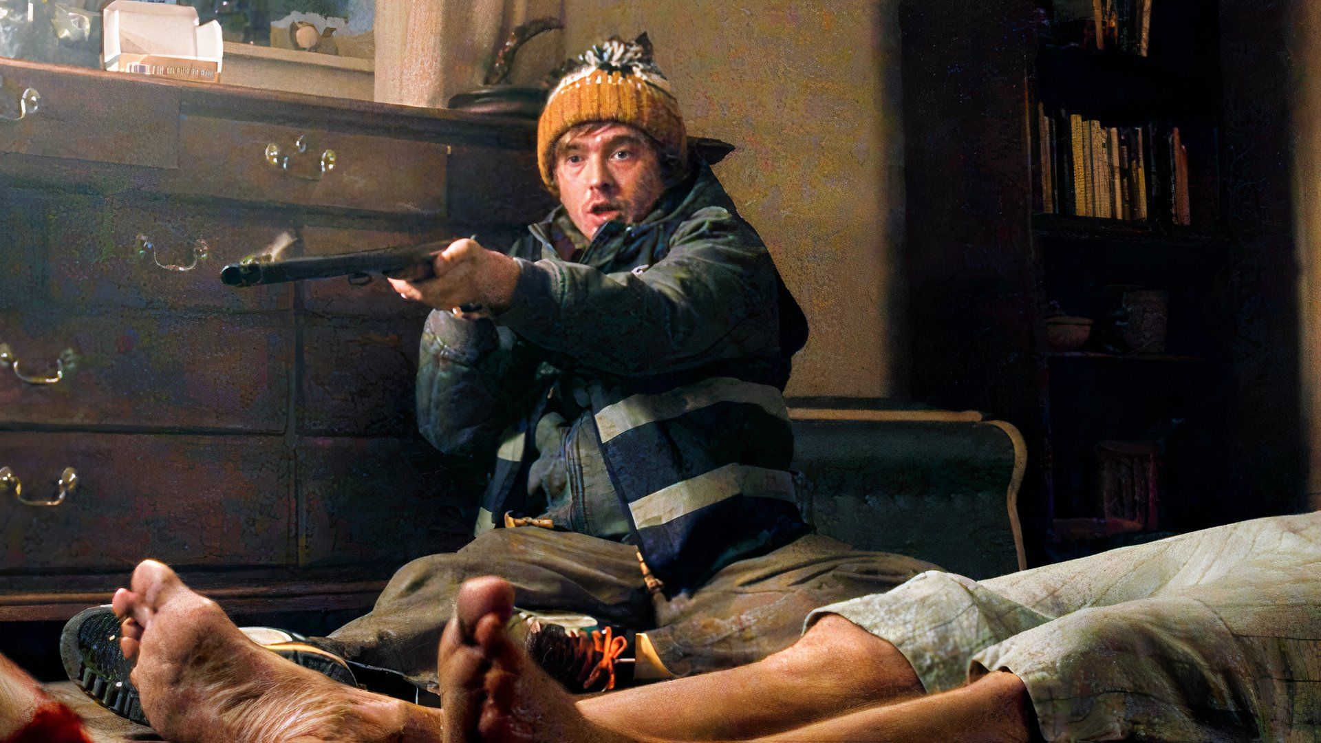 Austin Abrams as Bryan wearing a snow jacket and knitted cap, aiming a double-barrelled shotgun offscreen while sitting on the floor next to a body