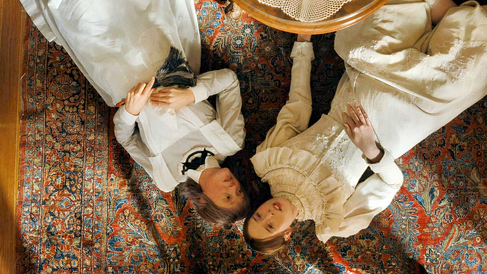 Vicky Krieps laying next to Ella Beatty on a colorful floor rug in white Victorian-style dresses