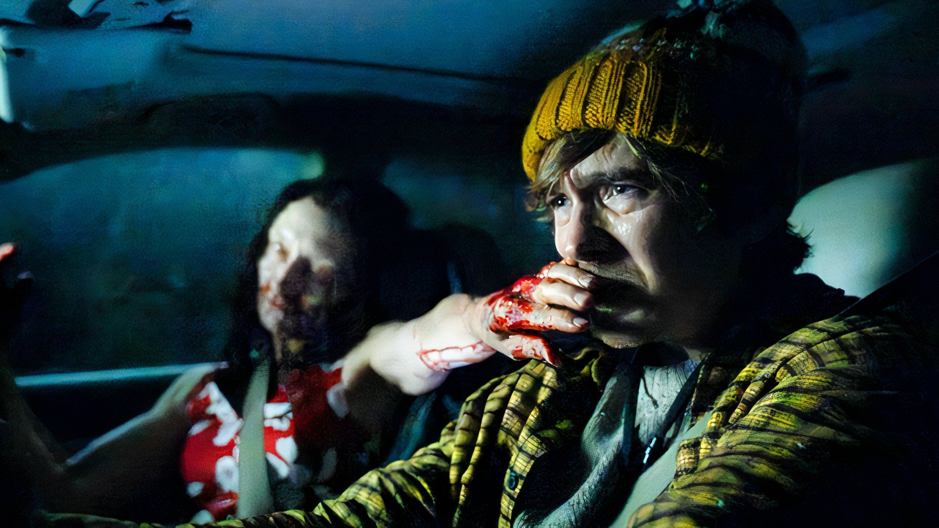 Austin Abrams as Bryan wearing a dark green and orange knitted hat and yellow flannel shirt, driving his car in a panic as a lady zombie reaches her bloodied hand over his mouth