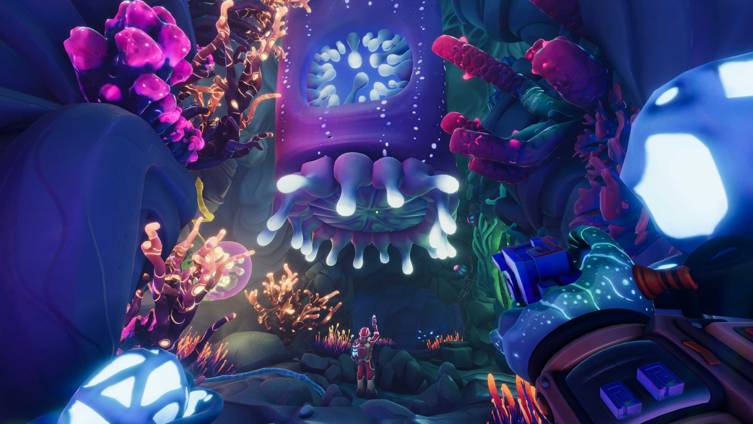 A red-suited Explorer stands beneath a massive purple alien organism with glowing white tendrils inside a cavern filled with colorful bioluminescent plants in We Were Here Tomorrow