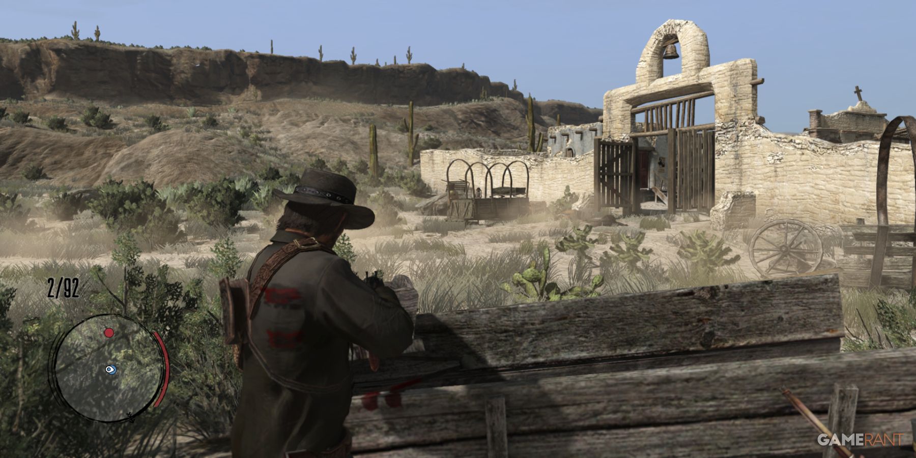 John Marston aiming from cover in a desert outpost in Red Dead Redemption, with a ruined fort visible