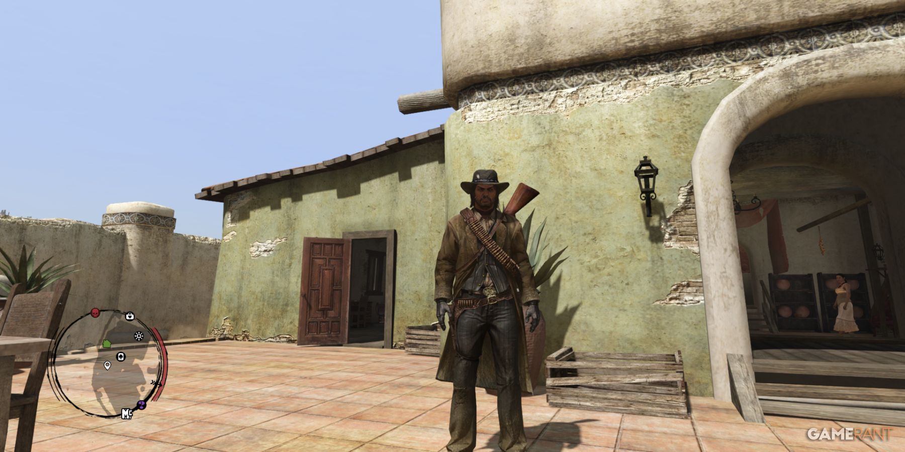 John Marston standing outside a rustic building with his rifle in Red Dead Redemption's arid landscape